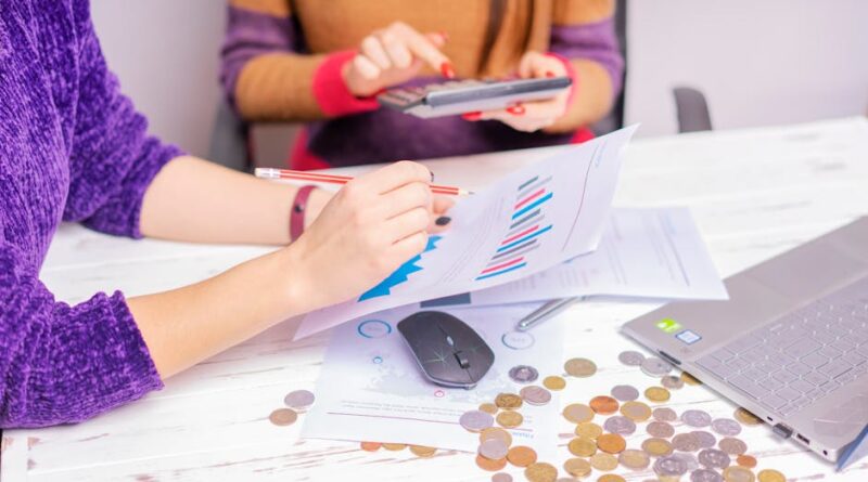 Two women analyzing financial charts with a laptop and coins on desk in an office setting.