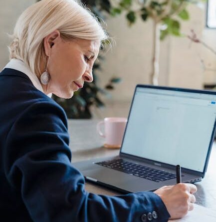 Elderly woman working on a laptop at office desk, writing notes with focus.